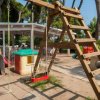 Area giochi con altalene, scivolo in plastica e tavolo da ping-pong tra gli alberi.