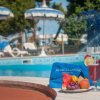Bordo piscina con piatto di frutta, bevanda e asciugamano blu. Ombrelloni sullo sfondo.