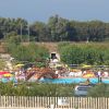 Vista di una piscina all'aperto con ombrelloni gialli e un ponte in legno, alberi e siepi intorno.