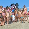 Un gruppo di bambini in spiaggia intorno a una mascotte sorridente.