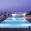 Piscina illuminata al tramonto, sdraio intorno e mare sullo sfondo.